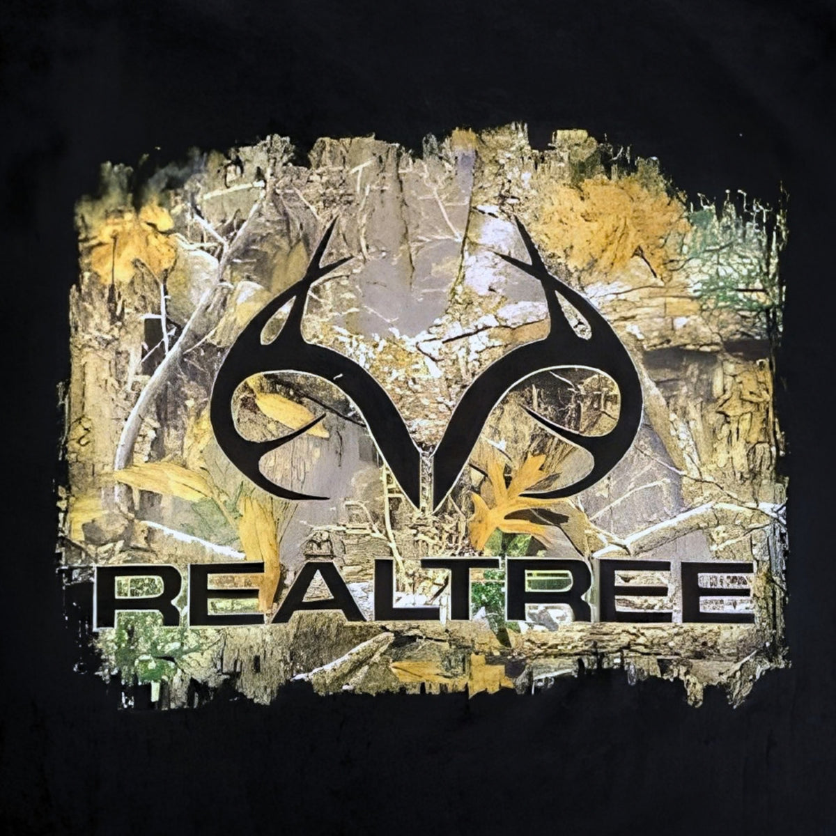 A close-up of a black garment featuring a large Realtree camouflage graphic on the chest. The graphic includes the black Realtree antler logo above the Realtree wordmark, set against a background of tan, green, and brown woodland camouflage with a distressed, torn-edge effect.