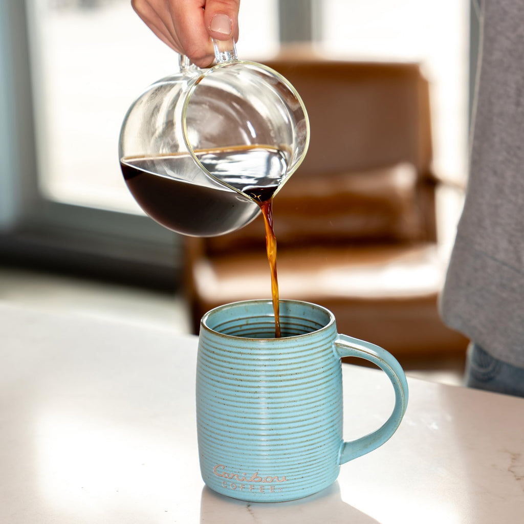 Person pouring coffee from a clear glass pot into a blue mug on a light surface.